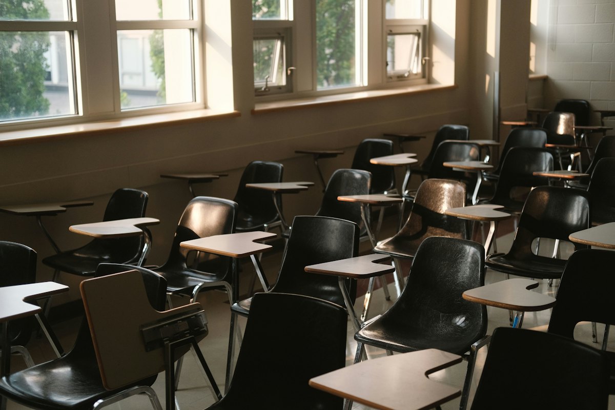 An empty classroom with desks and chairs beside tall windows